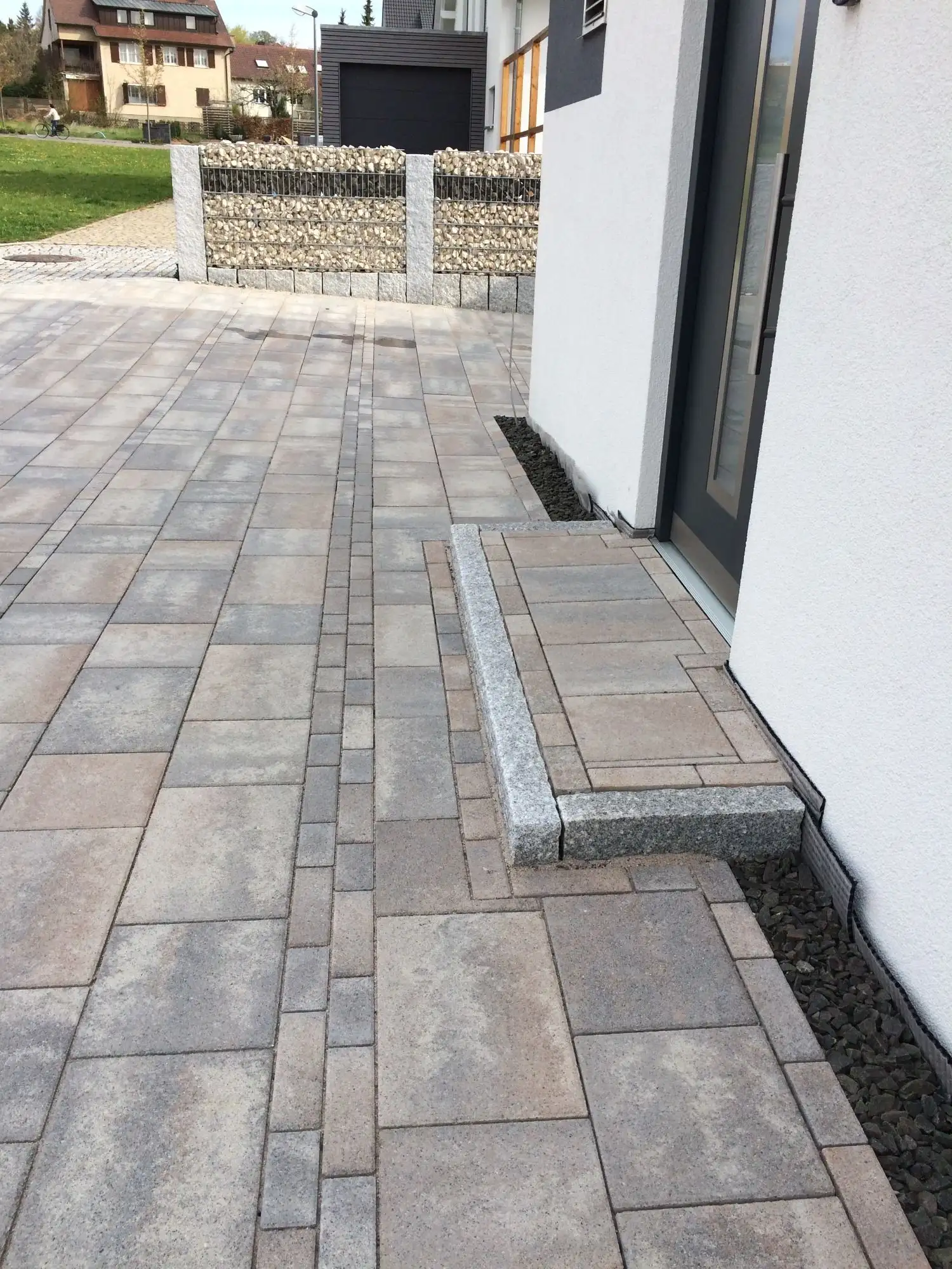 Hauszugang aus mehrfarbigem Rechteckpflaster mit Granit-Blockstufe, dahinter eine Gabione