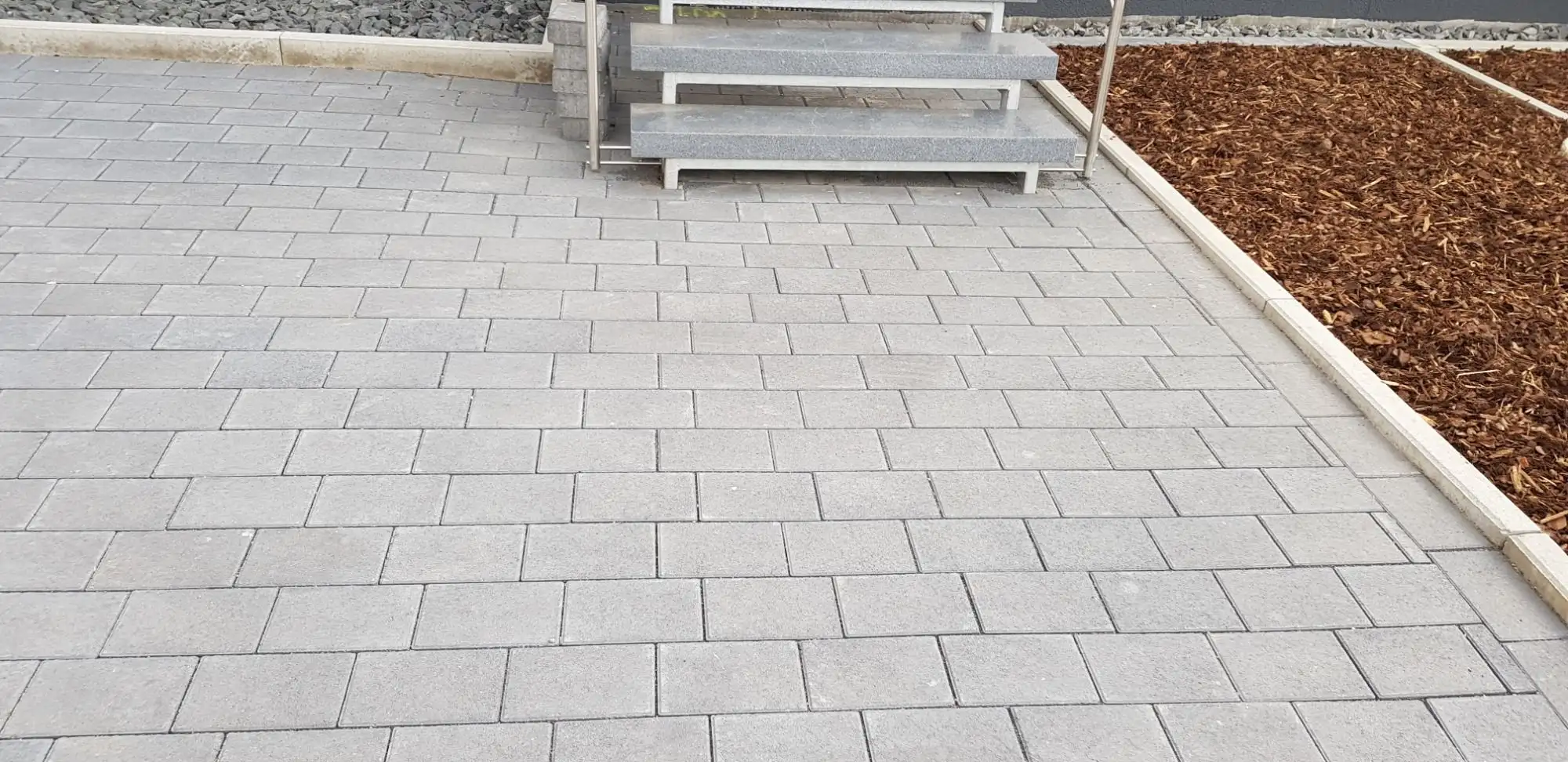 Hellgraue Pflasterfläche mit Blockstufen und Böschung mit Rindenmulch