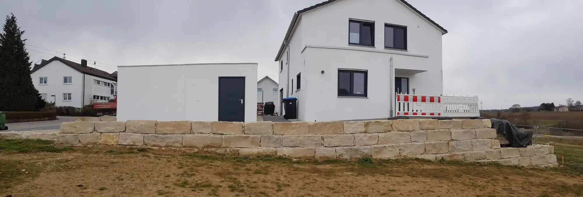 Kalkstein-Blockmauer an einem Neubau, davor unbefestigte Fläche
