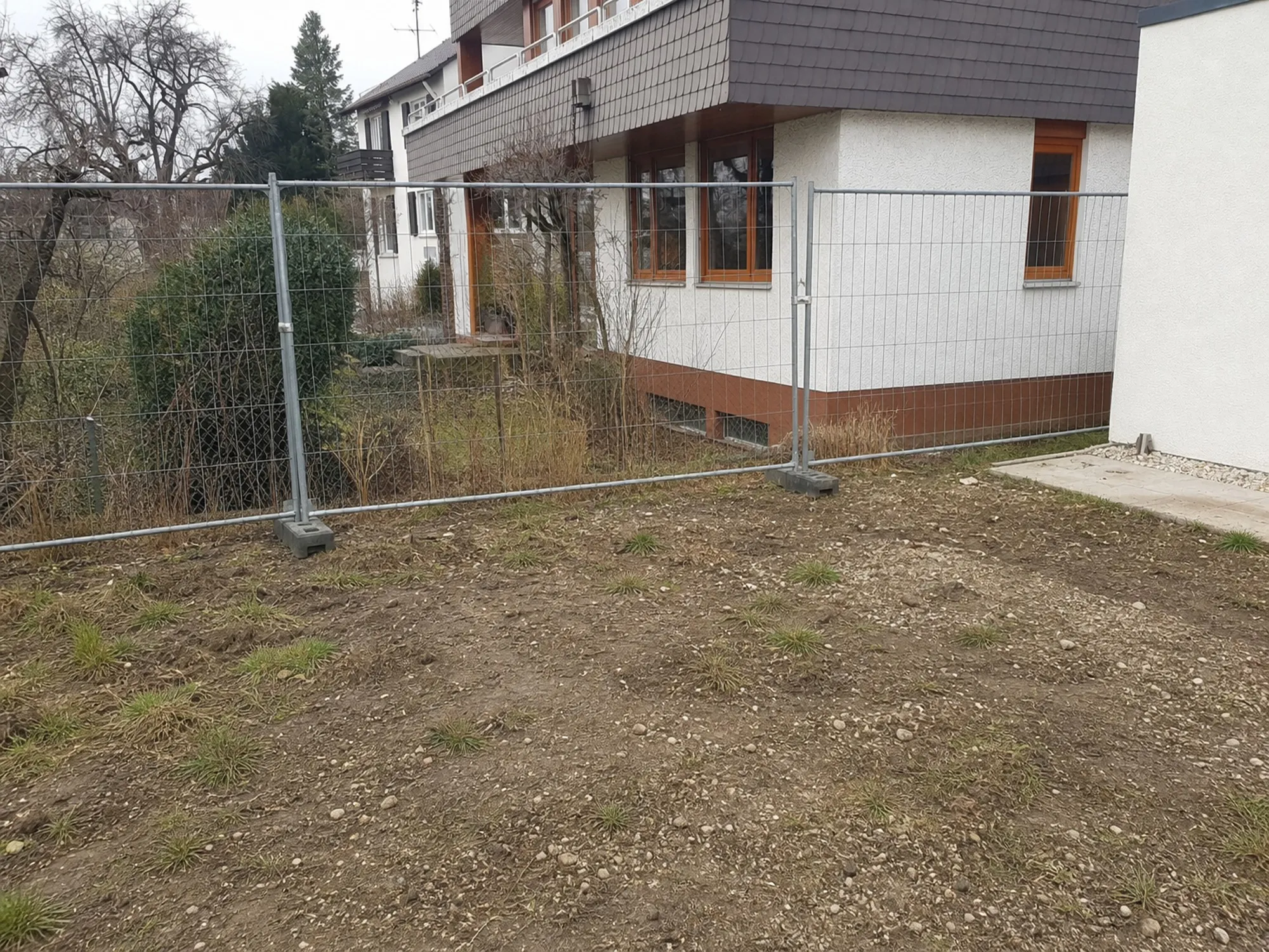 Nachgestellter Ausgangszustand: unebene Erde mit Grasbüscheln und Bauzaun vor dem Haus